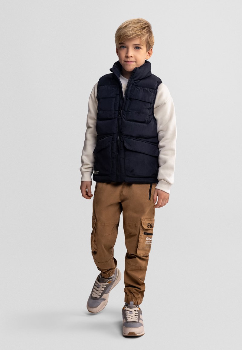 Garçon aux cheveux blonds portant une doudoune noire, un sweat-shirt beige, un pantalon cargo marron et des baskets grises, debout devant un fond clair.