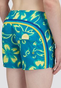 Man in blauwe en gele bloemen zwembroek met een achterzak en gebogen strepen aan de zijkant.