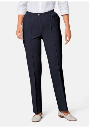Pantalons en tissu bleu marine avec taille élastique, plis sur le devant et texture lisse, associés à des mocassins clairs. Présente un détail de bouton au niveau de la taille.
