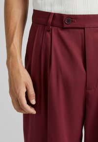 Pantaloni su misura bordeaux con pieghe, cintura in tessuto e chiusura con bottone. Texture liscia e design a gamba dritta.
