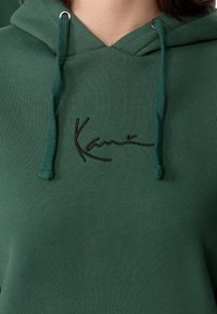 Sweat à capuche vert avec un cordon de serrage, des poignets côtelés et un logo noir brodé sur la poitrine. Texture douce avec un design sans couture.