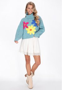 Tyrkysový pletený sveter s oversized strihom, zdobený viacfarebnými kvietkovými aplikáciami. Skombinovaný s bielou sukňou s čipkovým lemom a béžovými členkovými čižmami.