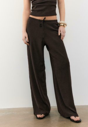 Kobieta ubrana w luźne ciemnobrązowe spodnie z prążkowanej tkaniny ze sznurkiem w pasie, dopasowany krótki top, sandały oraz duże bransoletki na prawej ręce.