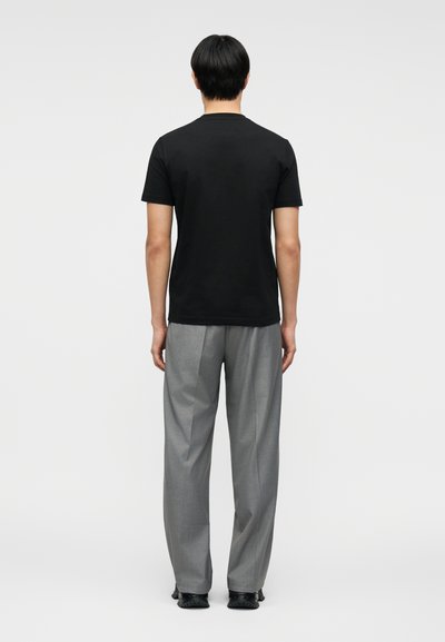 Άτομο που στέκεται με την πλάτη μας, φορώντας μαύρο πουκάμισο με κοντά μανίκια, γκρι παντελόνι και μαύρα παπούτσια, μπροστά σε ένα απλό φωτεινό φόντο.