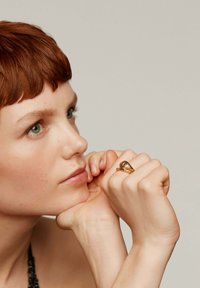 Anillo de oro con un diseño triangular y una piedra central, con una banda texturizada. El modelo muestra el anillo en una mano resting sobre su barbilla.