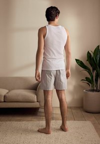 Weißes Tanktop und gestreifte Shorts in hellgrauen und pastellfarbenen Tönen. Steht barfuß auf einem Holzboden, mit einer Couch und einem Topf mit Pflanze in der Nähe.