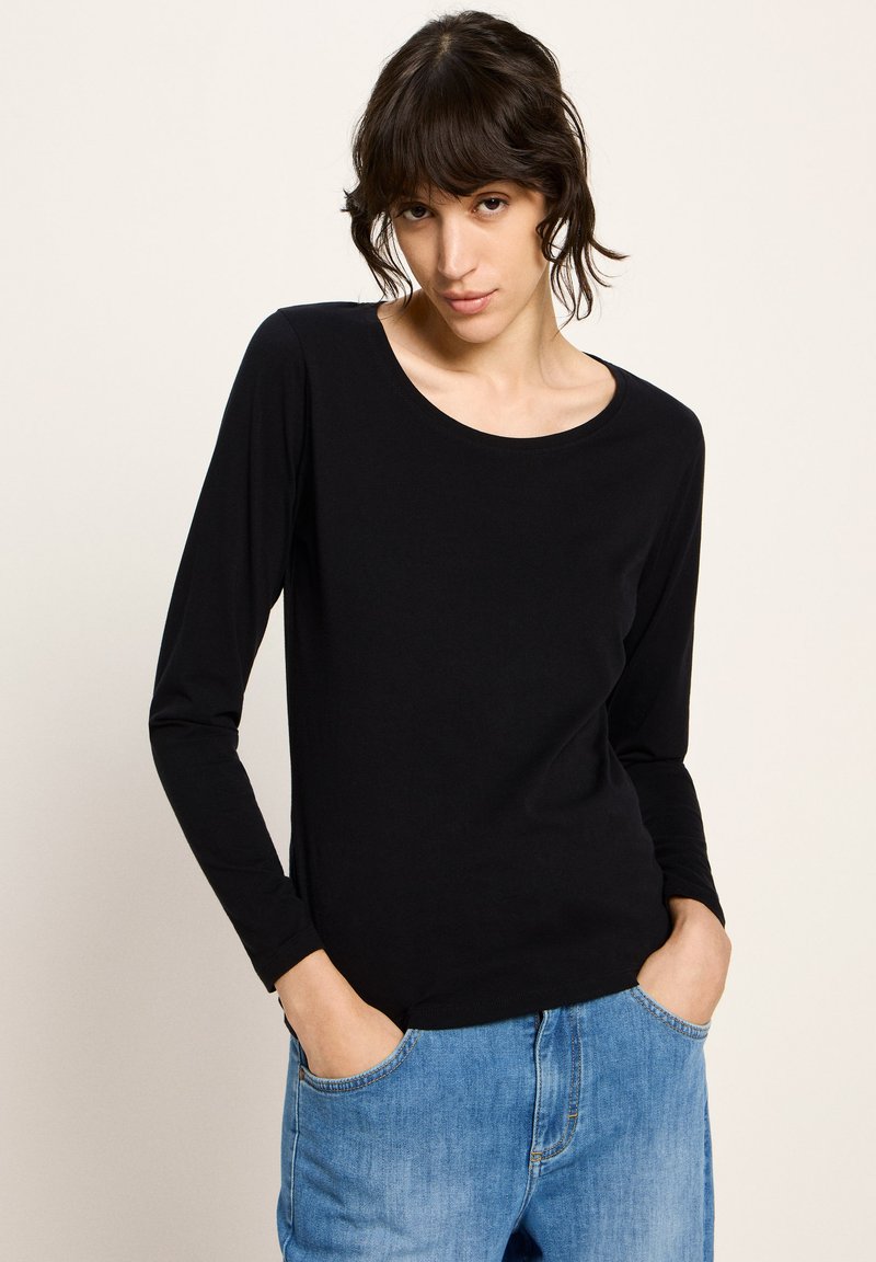 Frau mit dunklen Haaren, die ein schwarzes langärmliges Shirt und Bluejeans trägt, mit den Händen in den Taschen, vor einem schlichten hellen Hintergrund stehend.