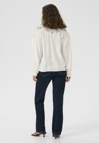 Blusa bianca a maniche lunghe con dettagli in crochet sulle spalle, abbinata a jeans blu scuro e sandali neri con tacco.