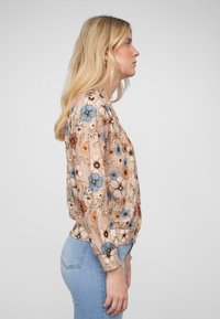 Blouse florale beige avec des fleurs bleues et orange, manches longues et poignets élastiques ; le tissu semble léger et texturé.