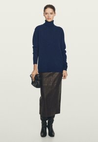 Donna che indossa un maglione a collo alto blu navy, gonna midi in pelle marrone scuro, stivali neri al ginocchio, che tiene una piccola borsa nera.