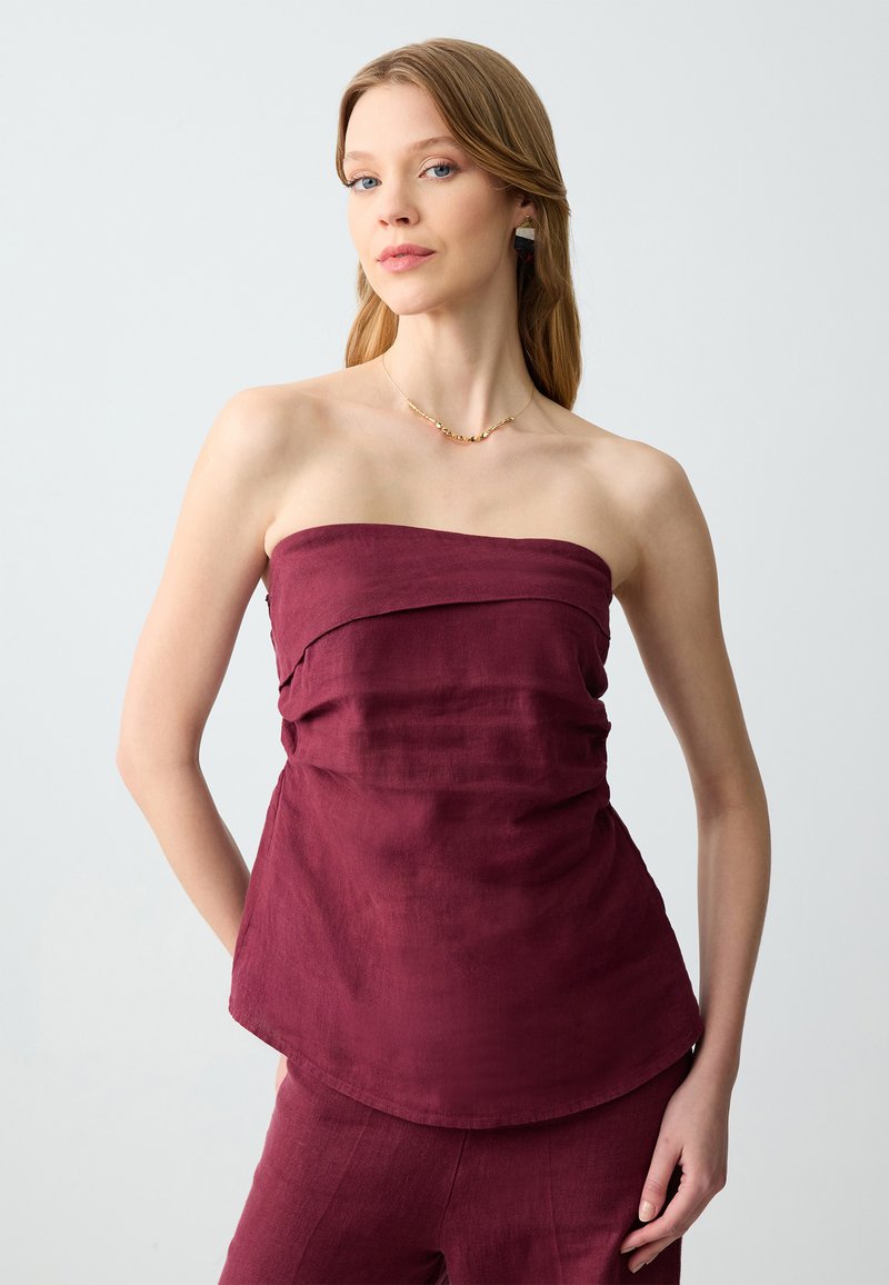 Top senza maniche color bordeaux in tessuto strutturato, con design drappeggiato e orlo arrotondato. Abbinato a gioielli dorati e uno sfondo morbido.