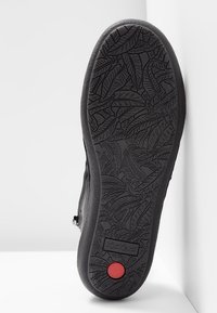 Semelle de chaussure noire avec un motif de feuille en relief, détail circulaire rouge près du talon, et le nom de la marque "Kickers" embossé au centre de la semelle.