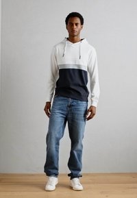 Felpa con cappuccio di colore bianco nella parte superiore, striscia grigia e parte inferiore blu navy, abbinata a jeans blu e sneakers bianche, su un pavimento di legno.