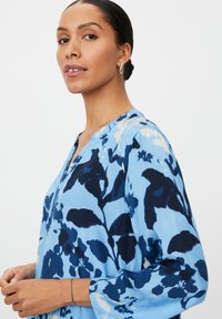 Bluzka w jasnoniebieskim kolorze z kwiatowym wzorem i ciemnoniebieskimi akcentami, z dekoltem w serek i długimi rękawami. Miękka, lekka tkanina.