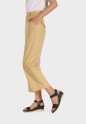 Donna con pantaloni beige corti e sandali neri con zeppa, in piedi con una mano in tasca davanti a uno sfondo semplice.