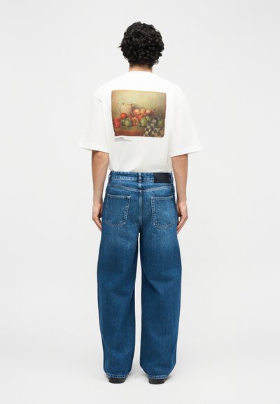 Personne aux cheveux bouclés portant un t-shirt blanc ample avec une impression de nature morte et un jean large en denim bleu, debout de dos.
