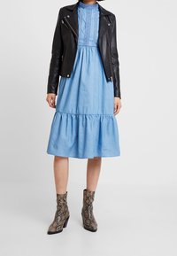Černá kožená biker bunda přes světle modrou šaty do délky kolen se zdobeným lemem, doplněná texturovanými kotníkovými botami s hadím vzorem.