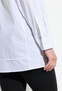 Witte langekousen shirt met een losse fit, voorzien van een knoopmanchet en subtiele stikdetails aan de zoom. Gecombineerd met zwarte spijkerbroek.