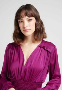 Blouse à col en V en satin magenta profond, avec des accents volants aux épaules et un bandeau smocké, dotée de manches longues.
