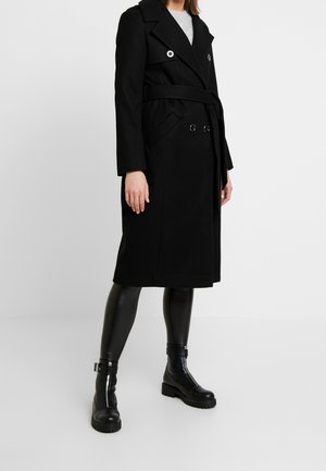 Femme portant un manteau noir ceinturé à double boutonnage, des leggings en cuir noirs et des bottines noires avec des détails de boucle, debout sur un sol blanc.