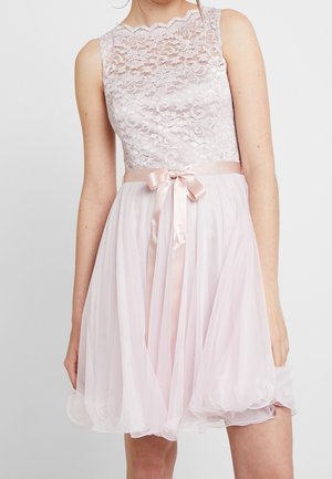 Ærmeløs lyserød kjole med blondekorsage, satinbånd bundet i taljen og flydende gennemsigtigt skørt, båret af person med afslappede arme.