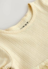 Ljusgul bomullstopp med ribbad textur, rund halsringning och fin spetskant längs fållen. Synlig märkning på insidan.