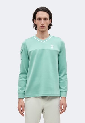 Homme aux cheveux foncés et ondulés portant une chemise à manches longues vert menthe avec un logo blanc et un pantalon clair, debout devant un fond uni.