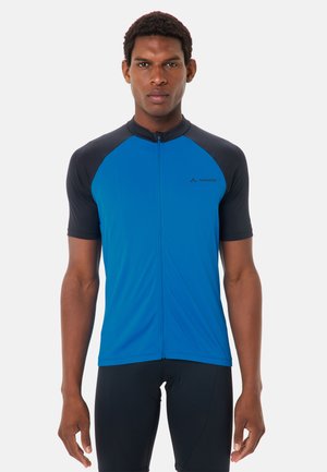 Uomo che indossa una maglia da ciclismo a maniche corte blu e nera con cerniera frontale e pantaloncini da ciclismo neri su sfondo bianco.
