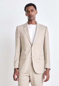 Beige Anzug mit einem einreihigen Sakko und passender Hose. Verfügt über einen strukturierten Stoff, zwei vordere Taschen und zwei Knöpfe. Darunter ein weißes Hemd.