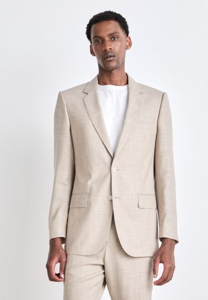 Beige Anzug mit einem einreihigen Sakko und passender Hose. Verfügt über einen strukturierten Stoff, zwei vordere Taschen und zwei Knöpfe. Darunter ein weißes Hemd.