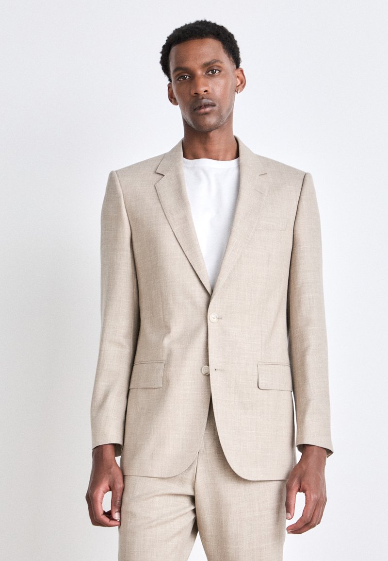 Beige Anzug mit einem einreihigen Sakko und passender Hose. Verfügt über einen strukturierten Stoff, zwei vordere Taschen und zwei Knöpfe. Darunter ein weißes Hemd.