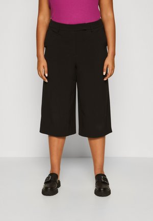Pantalons culotte noirs au design ajusté, présentant une texture lisse et des jambes larges. Associés à des mocassins noirs ornés de détails.