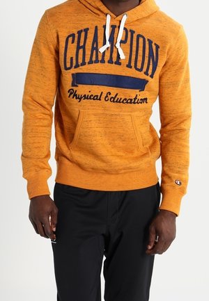 Oranger Hoodie aus strukturiertem Stoff, mit großem "Champion"-Logo in Marineblau, Kapuze mit Kordelzug und Vorderfach. Kombiniert mit einer schwarzen Hose.