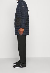 Marinblå puffjacka med quiltad textur, svart axelväska, svarta byxor och svarta sneakers med vita accenter.