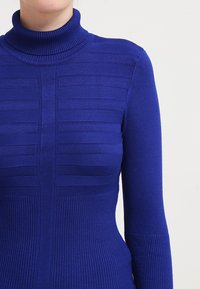 Blauer Rollkragenpullover mit gerippter Textur und horizontalen Streifenmustern, langen Ärmeln und tailliertem Design.