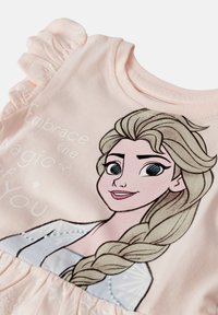 Jasnoróżowa koszula z krótkim rękawem, ozdobiona grafiką postaci z warkoczykami. Miękki materiał z falbaniastego wykończenia rękawów.