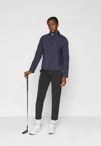 Veste navy avec une finition texturée, fermeture éclair à l'avant et poches latérales. Portée avec un pantalon noir et des baskets blanches, tenant un club de golf.