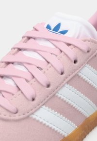 Close-up de um ténis em camurça rosa com riscas brancas, atacadores rosa, sola de borracha e um logo trefoil azul na língua branca.