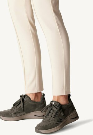 Person iført beige bukser og mørkegrå sporty sneakers med snørebånd og sidelommer med lynlås, stående på en lys baggrund.