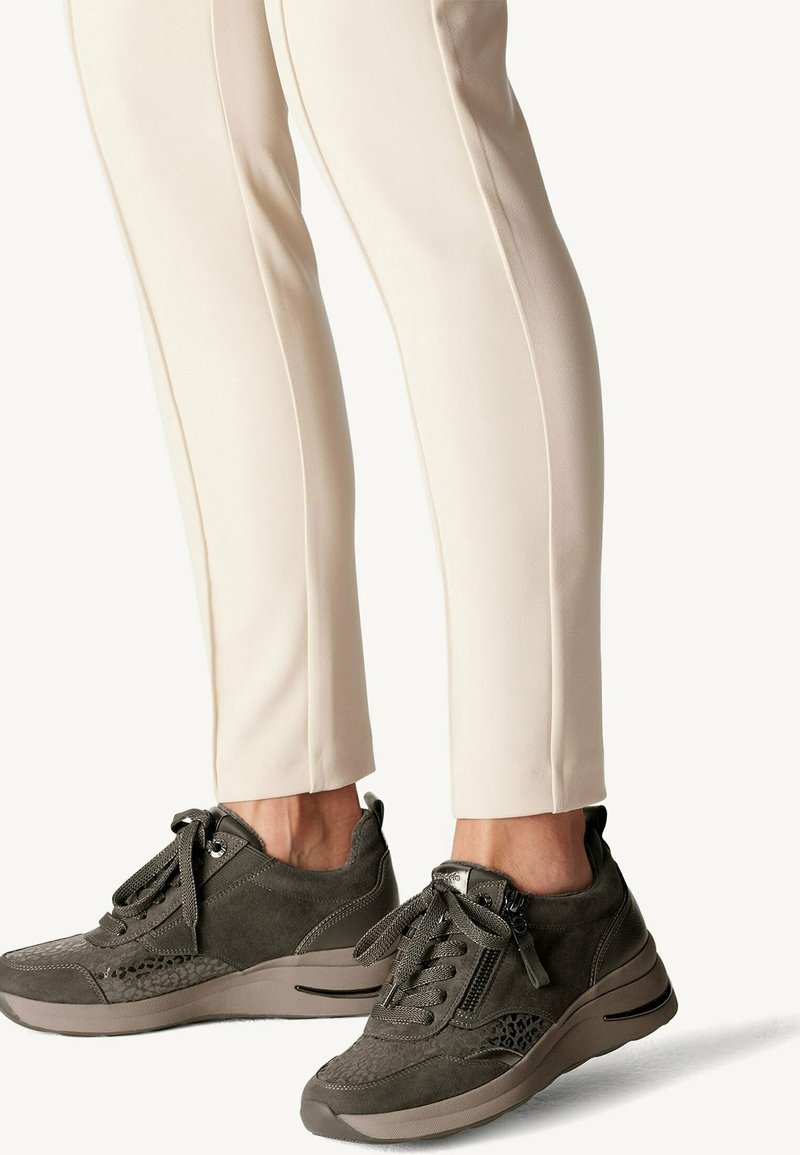 Personne portant un pantalon beige et des baskets sportives gris foncé avec lacets et fermetures éclair latérales, debout sur un fond clair.