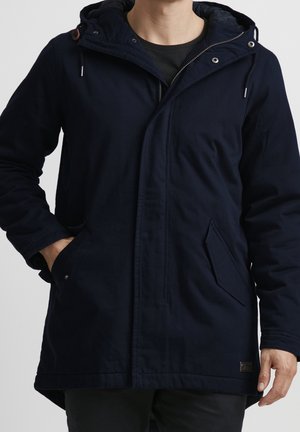 Homme portant une parka à capuche bleu marine avec fermeture éclair et boutons sur le devant, poches latérales, et pantalon noir, sur un fond uni.