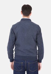 Giacca bomber blu in suede con colletto, polsini e vita a coste. Texture liscia, design pulito e vestibilità rilassata. Indossata sopra jeans scuri.