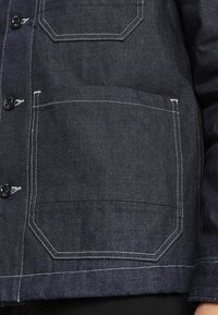 Tmavě modrá džínová bunda s kontrastním prošitím, dvěma velkými předními kapsami a zapínáním na knoflíky. Má strukturovaný design a texturovaný povrch.