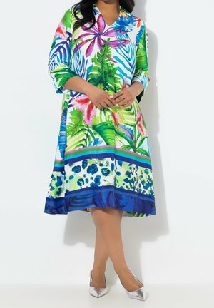 Femme portant une robe longueur genou, à manches longues, avec des motifs floraux tropicaux vibrants et des feuilles aux tons verts, bleus, roses et violets.