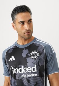 Camiseta de fútbol en gris y negro, hecha de material sintético, con un patrón de camuflaje, logo y texto de marca en blanco, y acentos a rayas en los hombros.