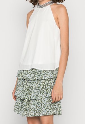 Witte mouwloze top met kralenomranding, gecombineerd met een groen en blauw gelaagde bloemenrok bij een persoon met krullend haar.