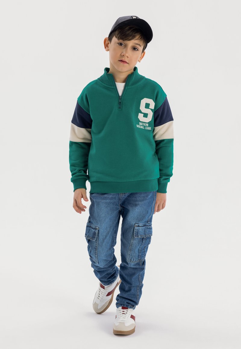 Felpa verde con zip, maniche blu e beige, con un logo bianco "S". Abbinata a jeans cargo azzurri e sneaker bianche.