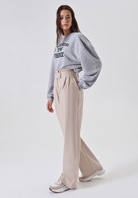 Donna che indossa pantaloni beige a vita alta e larghi, felpa grigia con scritta verde e sneakers bianche, in piedi contro uno sfondo neutro.