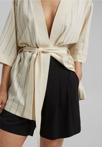Beige kimonomallinen yläosa, jossa pystysuorat raidat, syvä V-pääntie, leveät hihat ja solmittava vyö, yhdistettynä mustiin laskostettuihin shortseihin.
