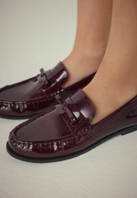 Bordeaux patentleren loafers met een verbonden strikdetail, moccasin-stiksel en een platte rubberen zool, close-up weergave.
